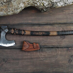 May include: A hand-forged axe with a black wooden handle and leather wrappings. The axe head is made of steel and has a hammered finish. The axe is accompanied by a brown leather sheath. The axe is engraved with the text "FEARLESS".