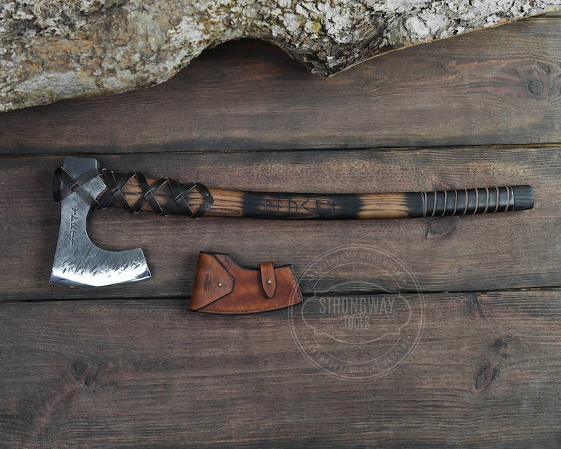 May include: A hand-forged axe with a black wooden handle and leather wrappings. The axe head is made of steel and has a hammered finish. The axe is accompanied by a brown leather sheath. The axe is engraved with the text "FEARLESS".