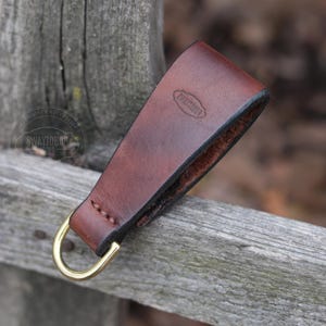 May include: A brown leather key fob with a brass ring. The fob has a stitched edge and the text "Swaytobog" is embossed on the back.