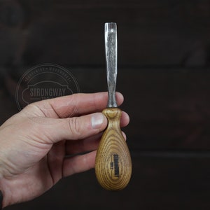 Puede incluir: Un formón de madera con un mango de madera marrón claro y una hoja plateada. El mango tiene el texto "STRONGWAY TOOLS" impreso. El formón se sostiene contra un fondo de madera marrón oscuro.