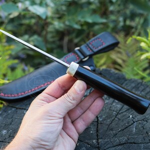 May include: A black-handled knife with a silver blade is held in a hand. The knife has a sheath with red stitching. The handle is dark and glossy, and the blade is sharp and pointed. The knife is resting on a wooden surface.
