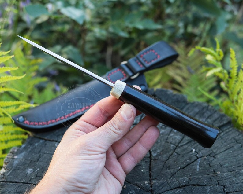 May include: A black-handled knife with a silver blade is held in a hand. The knife has a sheath with red stitching. The handle is dark and glossy, and the blade is sharp and pointed. The knife is resting on a wooden surface.