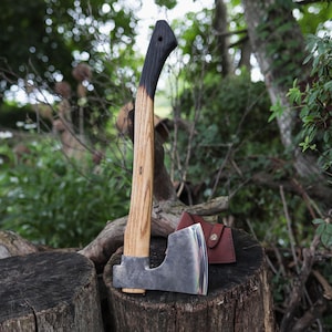 May include: A hand axe with a polished steel head and a long, curved wooden handle. The handle features a natural light wood grain finish that transitions into a dark, charred black colour at the top. A small brown leather sheath is included.