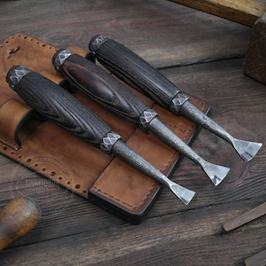 Puede incluir: Tres cinceles de talla de madera con mangos de madera negra y hojas de metal se muestran en un rollo de herramientas de cuero marrón. El rollo tiene un diseño cosido y el texto "SWAYTOOB TOOLS" es visible.