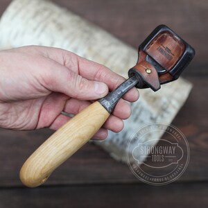 May include: A wood carving tool with a wooden handle and a metal blade. The tool is attached to a brown leather sheath with the text "ARMADOUR" embossed on it.