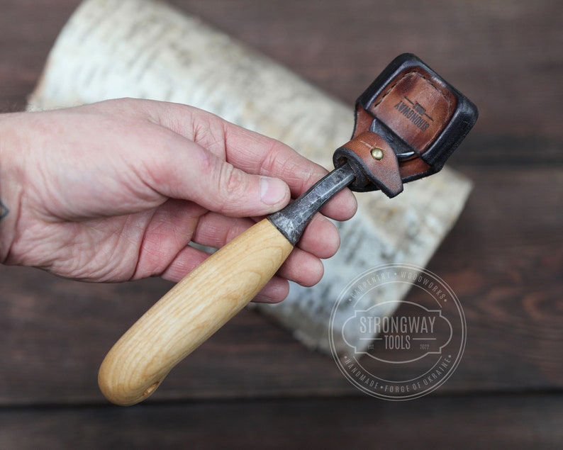 May include: A wood carving tool with a wooden handle and a metal blade. The tool is attached to a brown leather sheath with the text "ARMADOUR" embossed on it.