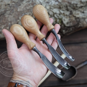 May include: Three wood handled carving tools with curved blades. The tools are held in a hand with a tattoo. The tools are made by Strongway Tools, which is a company that makes handmade forged tools.