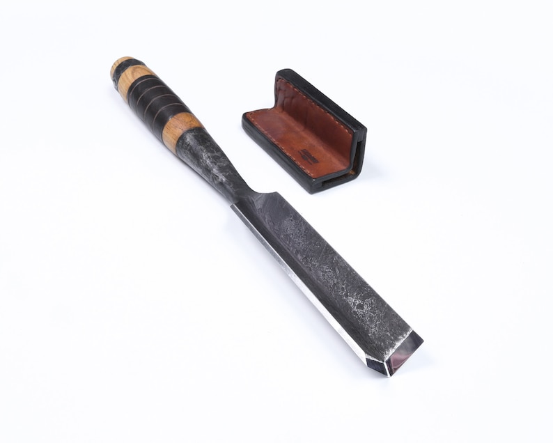 May include: A black and brown wood handled chisel with a leather sheath. The chisel has a beveled edge and a textured surface.