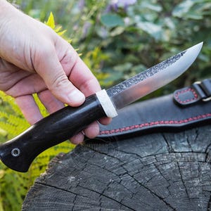 May include: A handcrafted knife with a dark wooden handle and a silver blade. The knife is held in a hand, with a black leather sheath with red stitching visible in the background. The knife appears to be a hunting or utility knife.