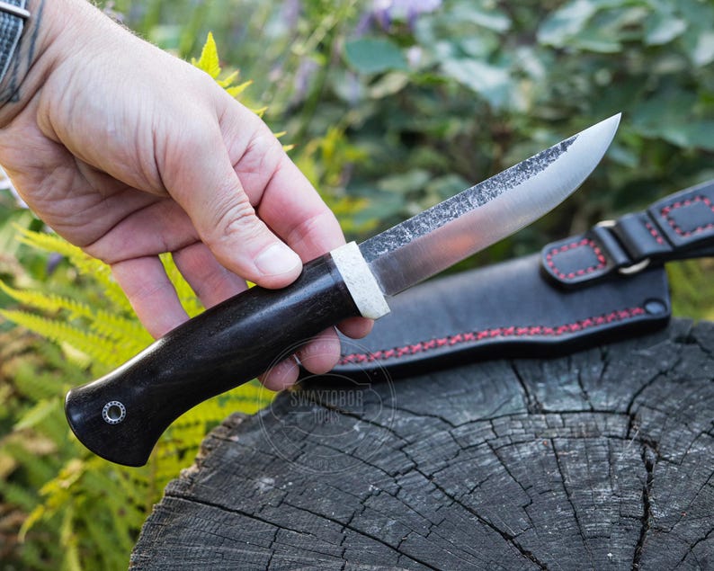 May include: A handcrafted knife with a dark wooden handle and a silver blade. The knife is held in a hand, with a black leather sheath with red stitching visible in the background. The knife appears to be a hunting or utility knife.