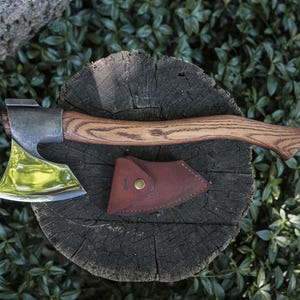 Puede incluir: Un hacha con una hoja de metal pulido y reflectante, un mango de madera curvado con veta visible y una funda de cuero marrón separada con un cierre de botón de presión de latón, sobre una superficie de madera oscura y circular.