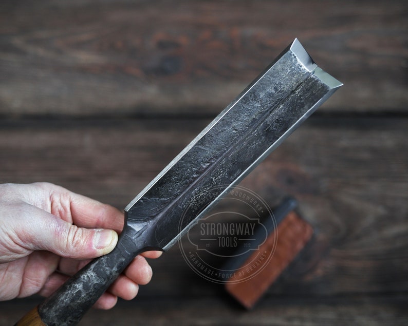 May include: A close-up of a hand holding a large, black metal chisel with a beveled edge. The chisel is marked with the text "Strongway Tools" and "Handmade - Forge of Celtic".