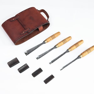 Puede incluir: Un juego de cuatro cinceles para tallar madera con mangos de madera y hojas de metal, junto con un estuche de herramientas de cuero marrón y cuatro protectores de hojas de cuero. Las herramientas están dispuestas sobre una superficie blanca.