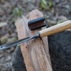 以下が含まれることがあります： 木製の柄と金属製の刃を持つノミが、木の板の上に置かれています。ノミは革製の鞘に入っています。