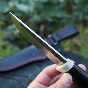 May include: Close-up of a handcrafted knife with a sharp, silver blade and a black handle. The handle has a white section near the blade. A black leather sheath with red stitching is in the background. The knife is held in a hand.