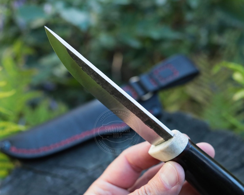 May include: Close-up of a handcrafted knife with a sharp, silver blade and a black handle. The handle has a white section near the blade. A black leather sheath with red stitching is in the background. The knife is held in a hand.
