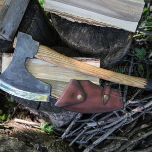 Può includere: Un'accetta con testa in metallo scuro dotata di lama curva e manico in legno marrone chiaro con estremità scura. Sotto la testa dell'ascia si trova un fodero in pelle marrone con due bottoni a pressione in metallo color ottone. Testo SWAYTOBOR TOOLS.