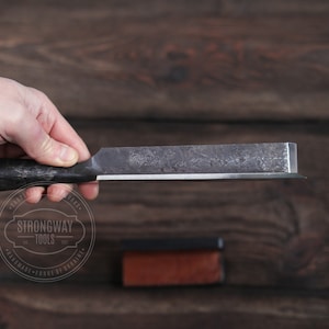 May include: A close-up of a metal chisel with a wooden handle. The chisel is held in a hand and the blade is sharp. The chisel is labeled "Strongway Tools - Hand-forged in Ukraine".