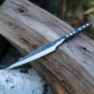 May include: A black and silver throwing knife with a pointed blade. The handle features white dot accents. The knife is displayed on a weathered wooden surface, highlighting its craftsmanship and design.