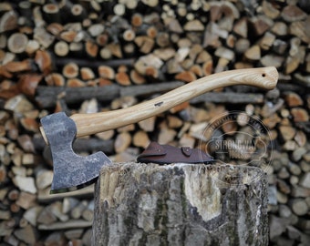 May include: A hand-forged axe with a wooden handle and a leather sheath. The axe is resting on a tree stump with a pile of firewood in the background. The axe is labeled "Strongway Tools".