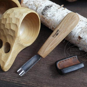 以下が含まれることがあります： 木製のハンドルと金属製の刃を持つ木彫りの道具。道具は革製の鞘と2つの木製のカップと一緒に木製の表面の上に置かれています。