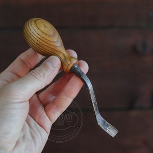 May include: A small, curved wood carving tool with a wooden handle. The tool is made of metal and has a sharp point. The handle is made of wood and has a brown and white striped pattern.