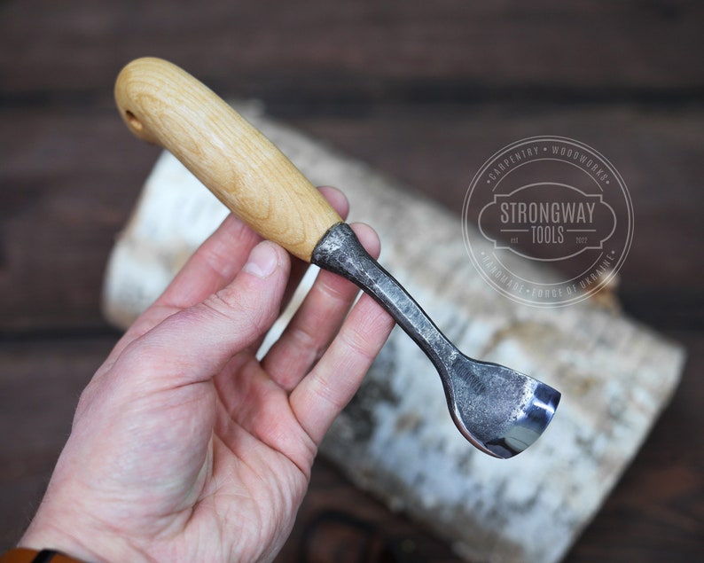 May include: A wood carving tool with a wooden handle and a metal blade. The blade is curved and has a sharp edge. The tool is held in a hand, and the background is a piece of birch bark. The tool is labeled "Strongway Tools".