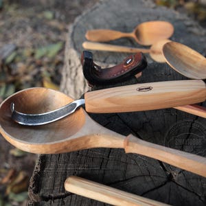 Puede incluir: Una cuchara de madera con un mango curvo y un gancho de metal. La cuchara está hecha de madera clara y tiene un acabado natural. El gancho está hecho de metal y está unido al mango de la cuchara. La cuchara está sentada sobre una superficie de madera. La cuchara es una herramienta para tallar madera.