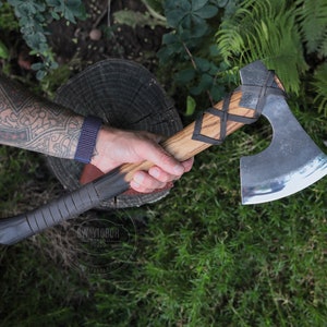 Könnte beinhalten: Eine Hand hält eine Axt mit schwarzem Griff, Lederwicklung und silberner Klinge. Die Axt hat einen Holzstiel mit Lederwicklung und silberner Klinge. Die Axt befindet sich auf einem grünen Hintergrund.