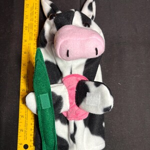 May include: A black and white cow hand puppet with a pink nose and a green felt accessory. The puppet is approximately 14 inches tall, with black spots and white patches. The puppet has a pink collar and a green felt accessory.