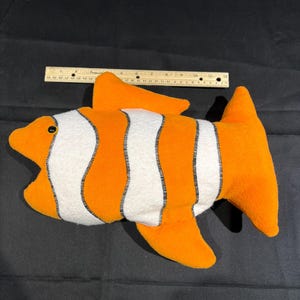 May include: A plush toy fish in orange and white, resembling a clownfish. The toy has black stitching and a black button eye. A ruler is placed next to the fish for size reference.