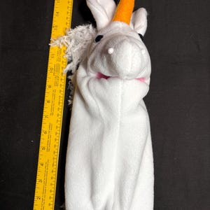 May include: A white plush unicorn hand puppet with an orange horn, black eyes, and a pink mouth. The puppet has white ears and a white mane. A yellow ruler is next to the puppet, measuring approximately 17 inches.