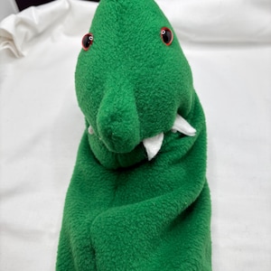May include: A green plush hand puppet shaped like a crocodile. The puppet has red eyes, white teeth, and a textured, fuzzy surface. The puppet's arms are folded, and the head is upright.