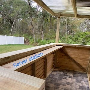 May include: A wooden outdoor bar with a serving station labeled "Server station". The bar is made of unfinished wood planks and has a corrugated metal roof. The bar is situated on a patio with a brick paver floor.