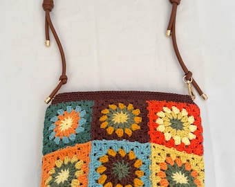 May include: A colorful crocheted bag with a brown leather strap. The bag is made of squares with different colors and patterns, including orange, yellow, green, blue, and brown.