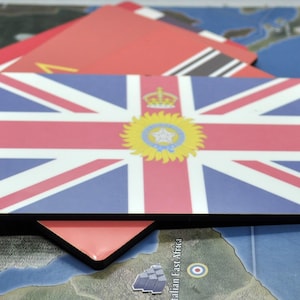 May include: Rectangular game pieces featuring flags of different nations. The primary piece displays the Union Jack with a central emblem. Other pieces show red and white flags, and a black and white flag. The pieces are on a map.