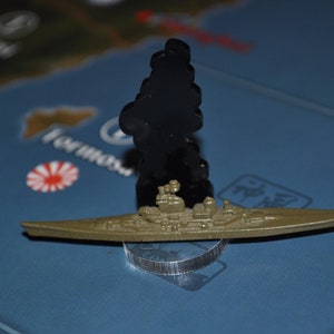 May include: A brown plastic model of a battleship with a black smoke plume above it. The model is sitting on a gray circular base.