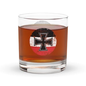 Puede incluir: Un vaso transparente lleno de un líquido marrón. El vaso presenta un gráfico circular desgastado de una cruz negra sobre un fondo blanco, con detalles en rojo y negro. El vaso se utiliza probablemente para servir bebidas.