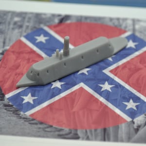 May include: A grey model of a Confederate ironclad warship sits on a red, white and blue Confederate flag. The flag is circular and has a white star in each corner.