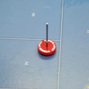 Peut inclure: Une pièce de jeu rouge avec une épingle argentée, portant l'inscription "Heavy Bomber" en lettres blanches.