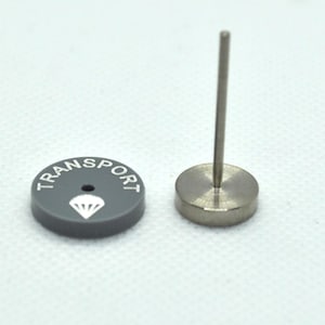 May include: A grey round button with the word "TRANSPORT" printed around the edge and a white parachute symbol in the centre. The button is attached to a silver metal post.