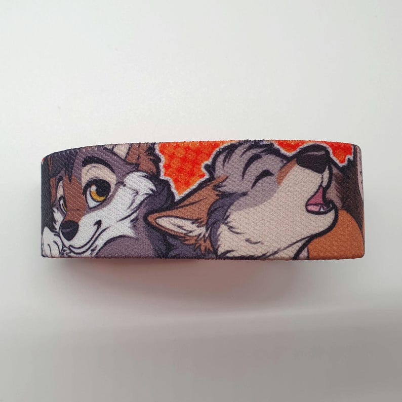 May include: A colorful wristband with a cartoon image of two wolves. The wolf on the left is smiling and the wolf on the right is howling. The background is orange with a pattern of small circles.
