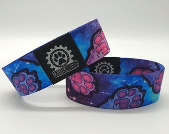 May include: Two blue and purple woven wristbands with a galaxy print and pink paw prints. The wristbands have a black patch with a silver gear and paw print logo that reads "Crafty Workshop".