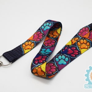 Pode incluir: Um cordão colorido com fundo preto e um padrão repetido de pegadas em várias cores, incluindo rosa, azul, laranja e amarelo. O cordão tem um clipe de metal prateado.