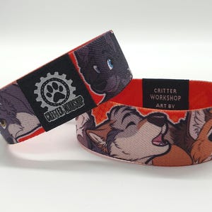 Peut inclure: Bracelet tissé rouge, blanc et gris avec un logo noir et gris qui dit "Critter Workshop Art By". Le bracelet présente une image de dessin animé de trois animaux à fourrure.