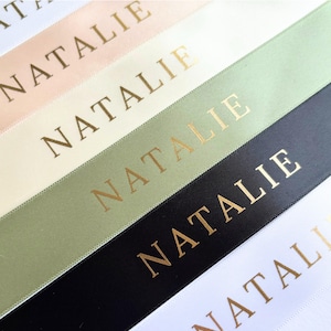 May include: Close-up of several ribbons in various colors, including white, blush, cream, sage green, and black. Each ribbon is printed with the name "NATALIE" in gold lettering. The ribbons are arranged diagonally.