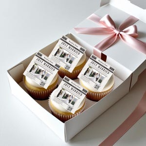 May include: A white gift box tied with a pink ribbon, containing four cupcakes. Each cupcake is topped with white frosting and a small paper with the text "JUST MARRIED" and a wedding photo. The cupcakes are in a white box.
