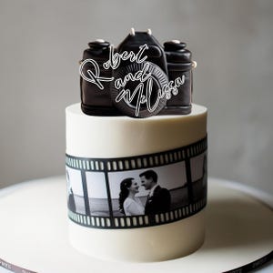 May include: A white wedding cake decorated with a black and white film strip border and a black camera topper with the text "Robert and Melissa" written in white.