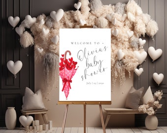May include: Welcome to Olivia's baby shower sign with a pink and white polka dot umbrella with red hearts. The sign says "Welcome to Olivia's baby shower" and "July 1st, 2019".