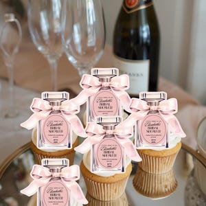 May include: Five cupcakes topped with white frosting and perfume bottle-shaped favors with pink bows. The favors read "Elizabeth's Bridal Shower". Champagne and wine glasses are in the background.